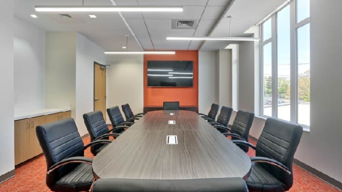 Interior conference room with oval table, leather seating, mounted television on orange wall, and windows.