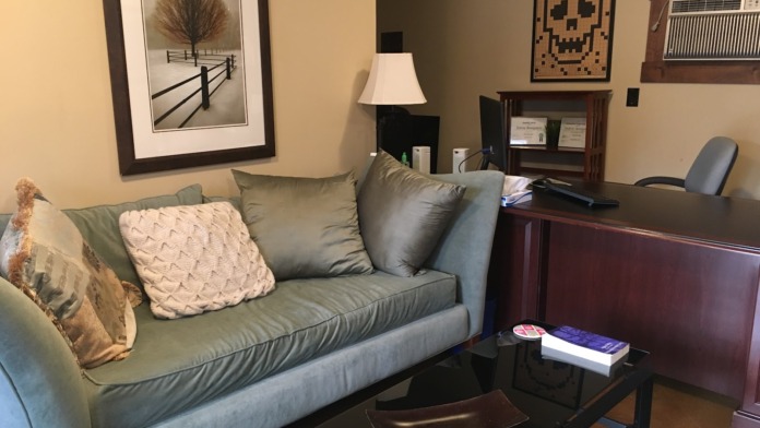 Interior lounge with gray sofa, decorative pillows, framed artwork, desk, shelving unit, and windows with outdoor view.