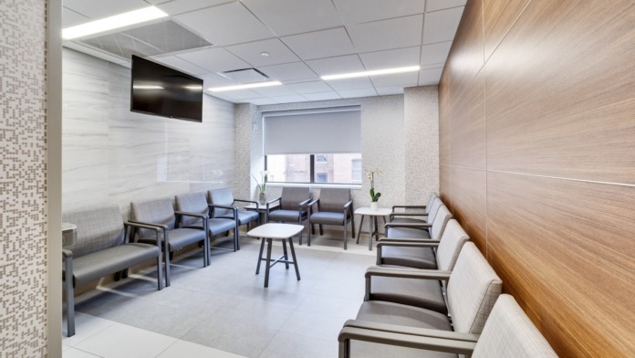 Modern waiting area with gray upholstered seating, wooden wall panels, white tile flooring, and windows.