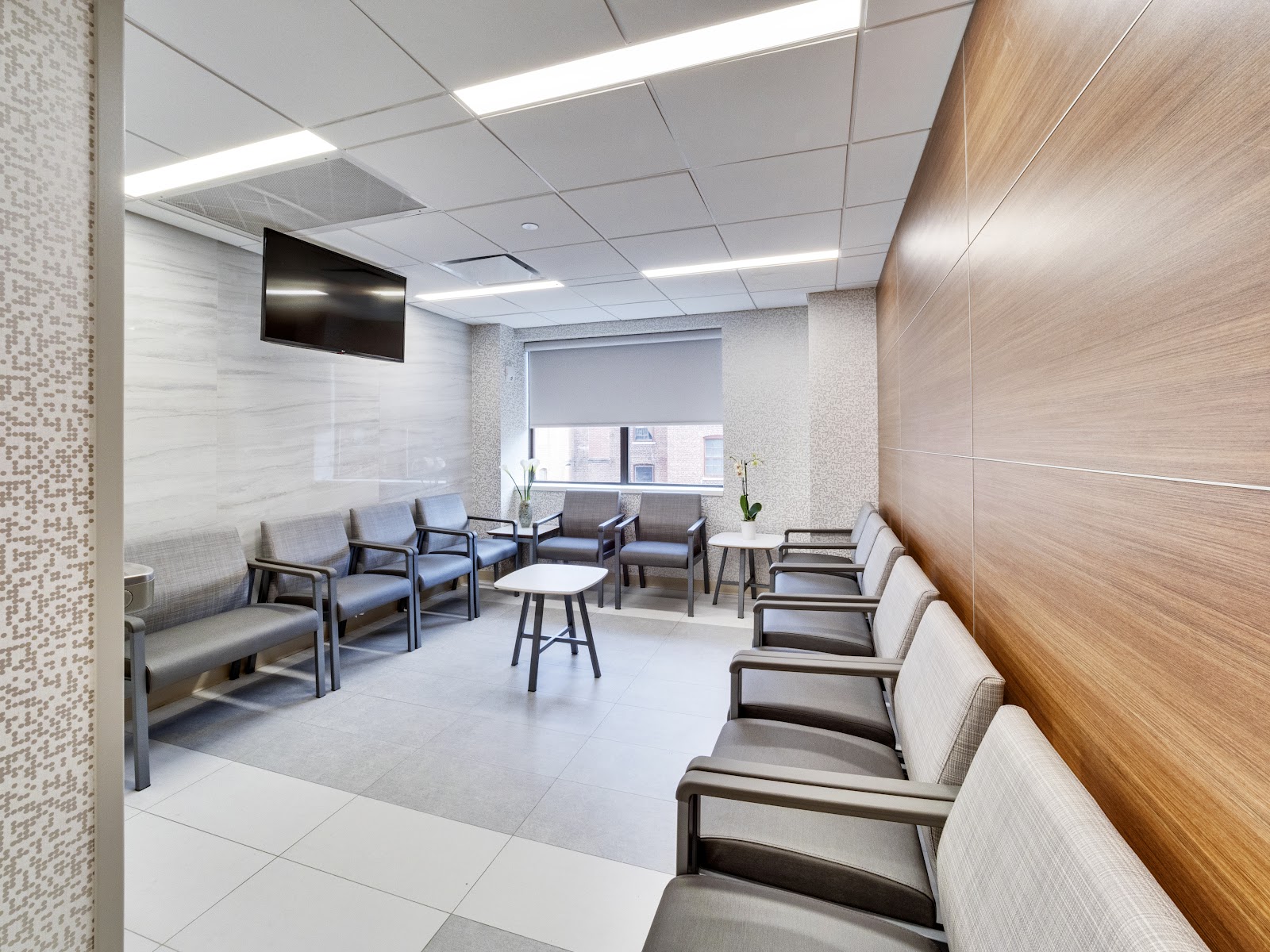 Modern waiting area with gray upholstered seating, wooden wall panels, white tile flooring, and windows.