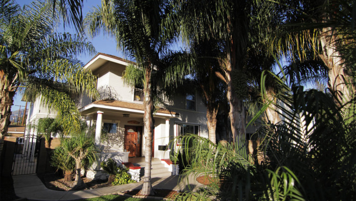 Two-story white building framed by mature palm trees, stone foundation, metal gates, and landscaped grounds.