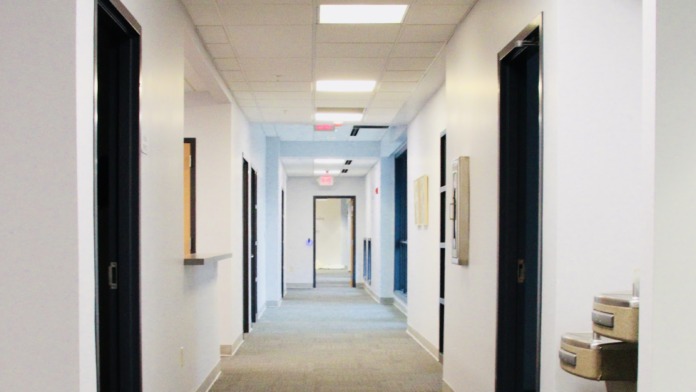 Interior hallway with multiple patient room doors, ceiling-mounted lights, white walls with blue accent stripe, carpeted flooring, and storage cubbies