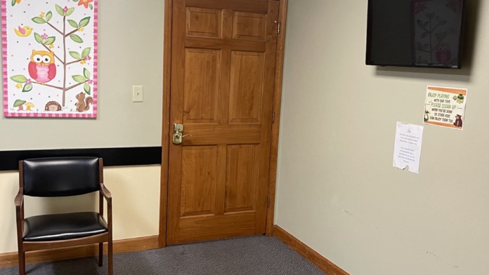 Interior room with beige walls, wooden door, wall clock, decorative poster, seating, and mounted television screen