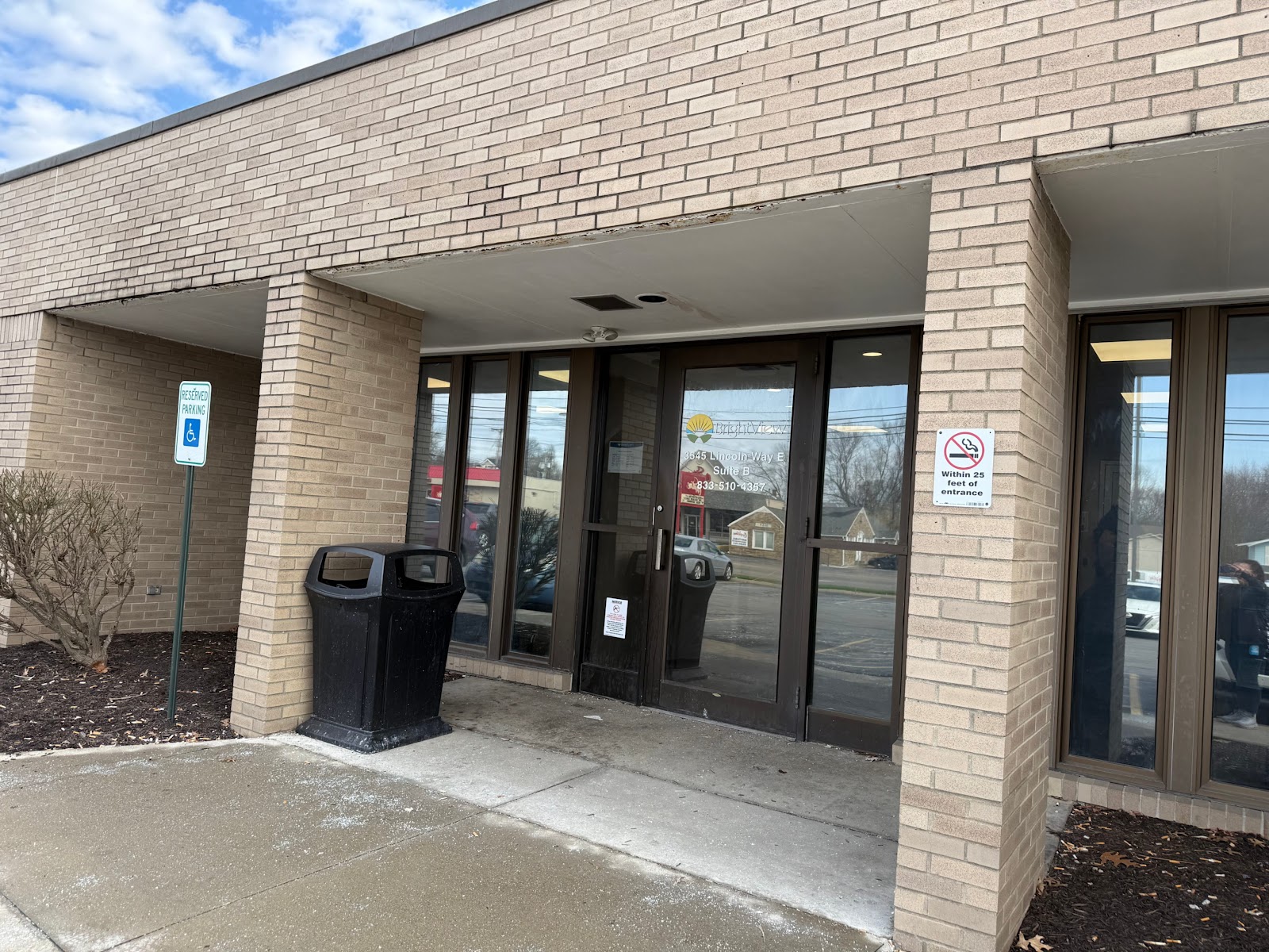 Exterior covered entrance with glass doors, brick walls, trash receptacles, and parking lot view.