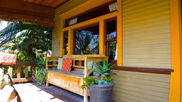 Exterior covered porch with yellow siding, wood beams, wooden bench seating, and potted plants