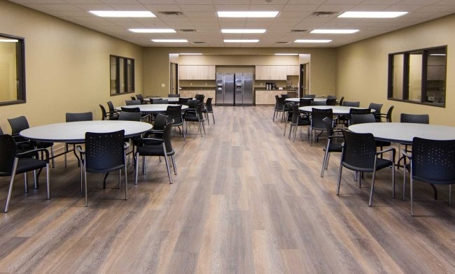 Interior dining room with long white tables, black chairs, equipped kitchen visible in background, wood flooring.