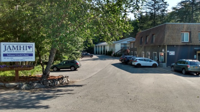 Exterior parking and driveway view with JAMHI signage, multiple vehicles, building structure, and forested setting.