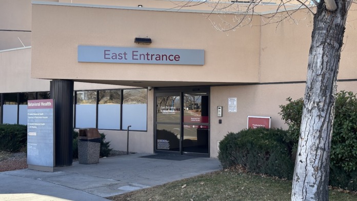 Exterior east entrance with glass doors, blue directional signage, tan building facade, and landscaping
