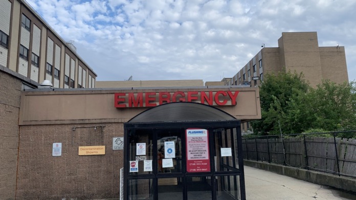 Emergency department entrance featuring red EMERGENCY signage, glass doors, brick facade, and posted notices.