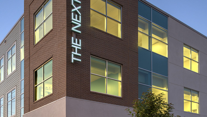 Exterior corner view of facility with illuminated 'THE NEXT DOOR' vertical signage and lit windows at dusk.