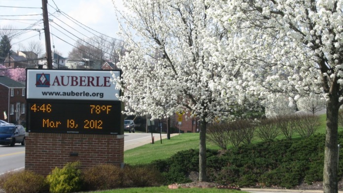 Facility entrance signage for Auberle with digital display, brick landscaping base, blooming trees, and building visible in background.