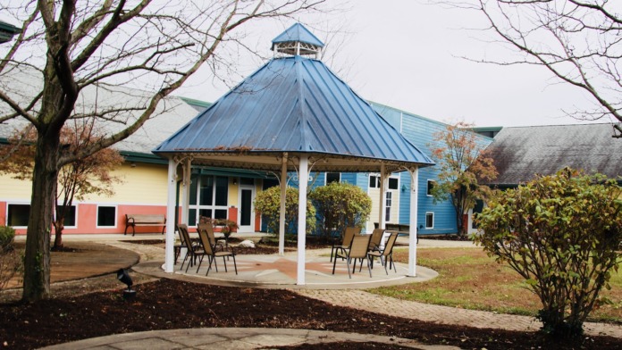 Exterior view of multicolored facility buildings with blue-roofed gazebo, mature trees, and patio seating area with landscaped grounds