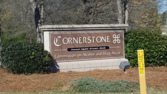 Exterior close-up of brown Cornerstone signage with facility logo and commission designation, surrounded by landscape plantings.