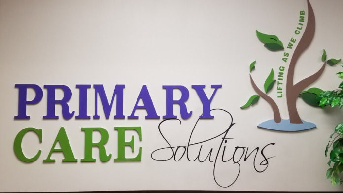 Interior signage showing Primary Care Solutions logo with green and blue tree graphic and tagline on white wall