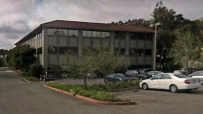 Exterior modern building with extensive windows, parking area with vehicles, and mature landscaping.