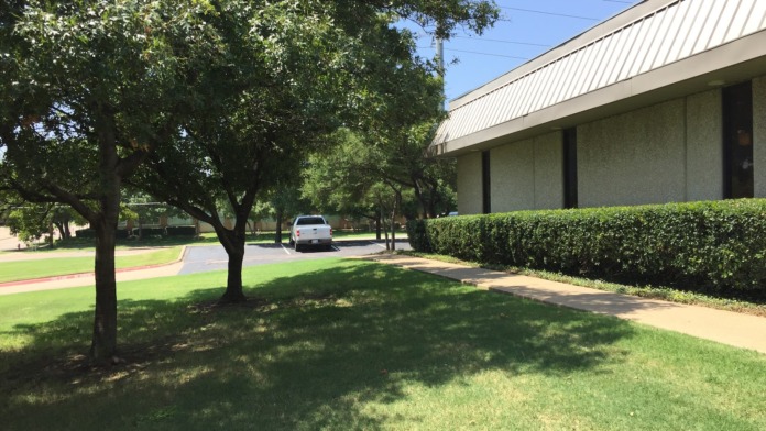 Exterior side view of building with mature shade trees, green lawn, tan facade, and metal roof overhang.