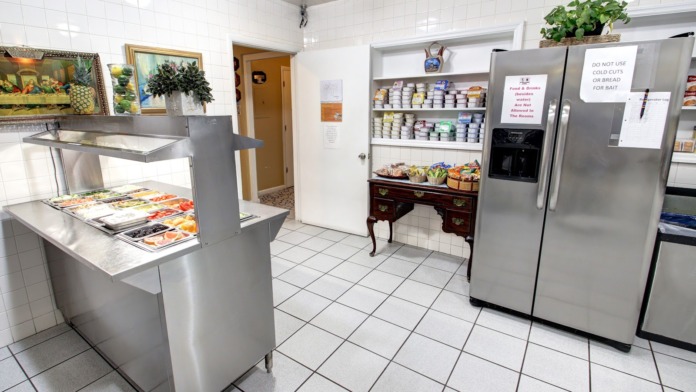Interior kitchen area with stainless steel refrigerators, white tile walls, food storage shelves, and serving counter displays