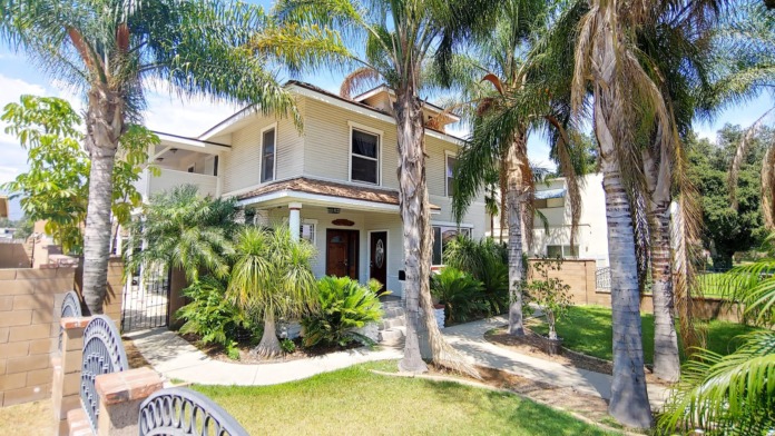 Two-story white residential building with manicured lawn, multiple palm trees, brick columns, and metal fencing.