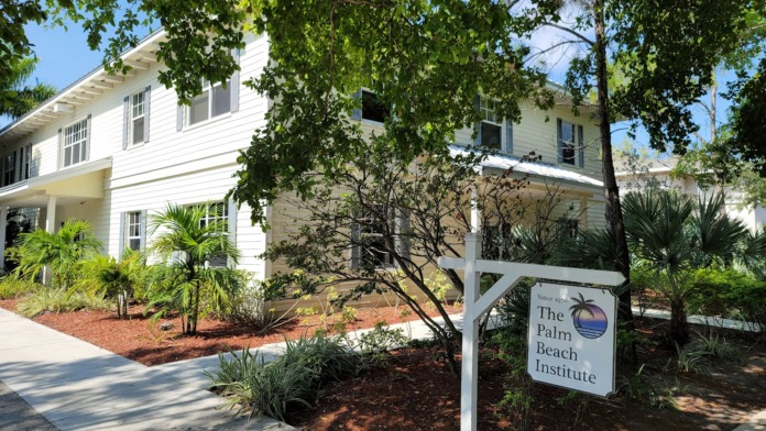 Exterior front facade of two-story white residential building with The Palm Beach Institute signage, tropical landscaping, brick walkway, and palm trees.