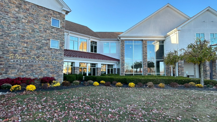 Modern facility front exterior showing brick and white architecture with flowering plant beds and clear grounds.