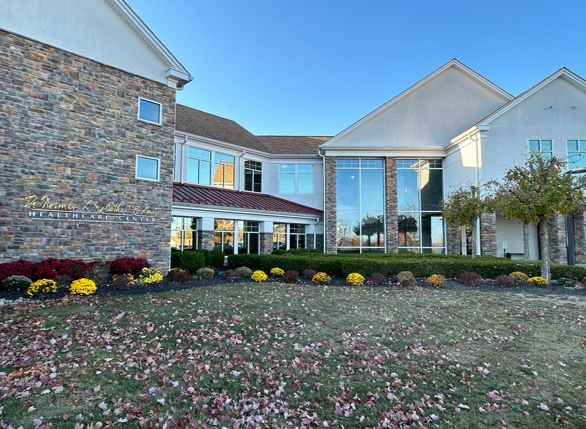 Modern facility front exterior showing brick and white architecture with flowering plant beds and clear grounds.