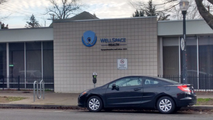 Exterior front facade of WellSpace Health showing tile walls, blue logo signage, and street-level entrance with parking