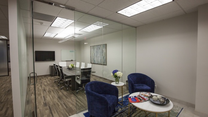 Interior office suite with glass-walled conference room, meeting table, blue upholstered chairs, and flowers