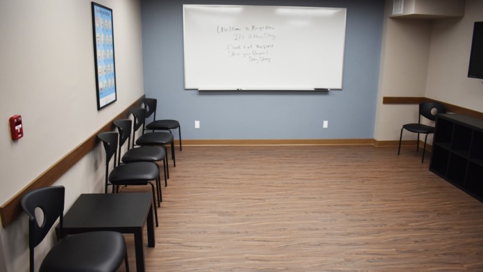 Interior room with blue-gray walls, whiteboard, wooden flooring, and arranged black chairs for group sessions.