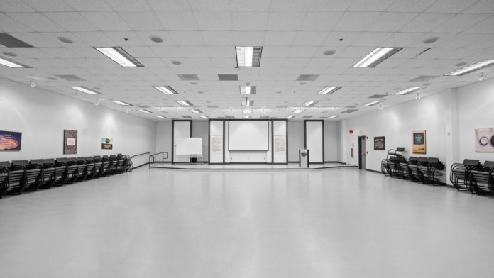 Interior large meeting space with white flooring, drop ceiling, glass-paneled dividers, and rows of black chairs for group gatherings
