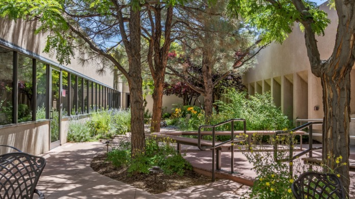 Covered outdoor courtyard with mature trees, metal benches, tables, windows, and landscaping throughout