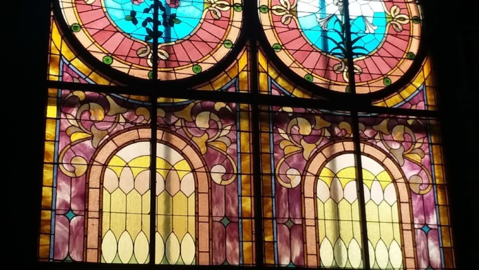 Interior stained glass window featuring symmetrical design with pink, blue, and yellow sections and religious cross symbols.