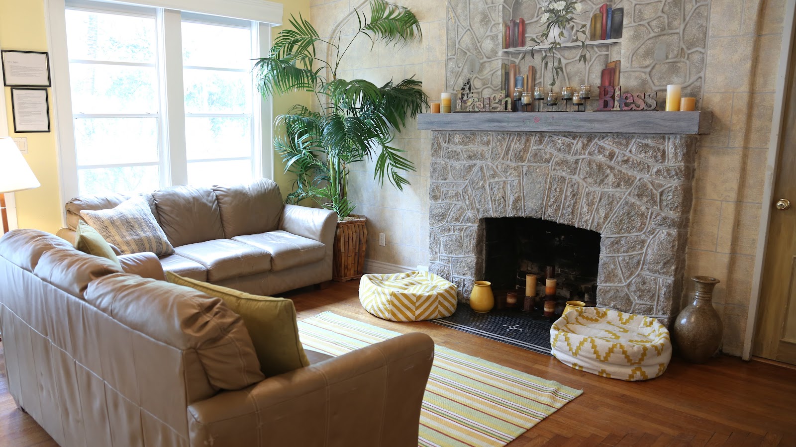 Interior living room with brown leather sectional sofa, stone fireplace with mantel, large windows, potted palm plant, and hardwood flooring.
