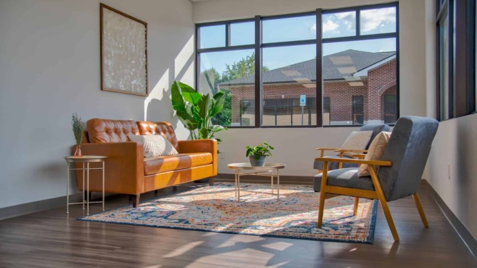 Interior lounge with cognac sofa, gray chair, potted plant, and large windows overlooking grounds