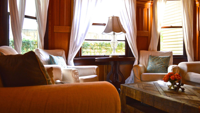 Interior lounge with upholstered seating, wooden side table with flowers, windows with sheer curtains
