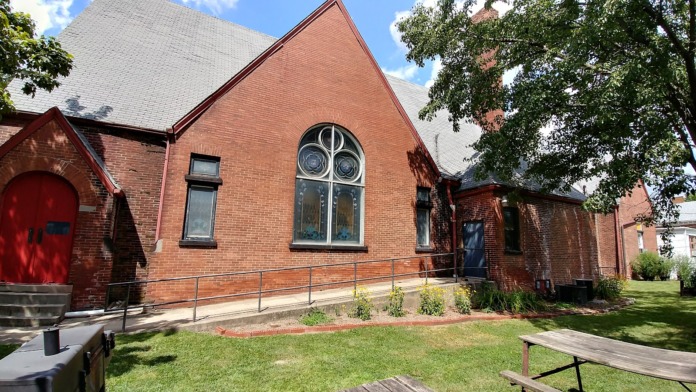 Exterior view of red brick building with large arched window, landscaped lawn, metal railing, and architectural details.