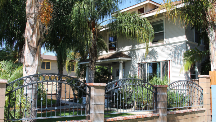 Two-story white residential building with mature palm trees, metal ornamental fencing, and green lawn from street view.