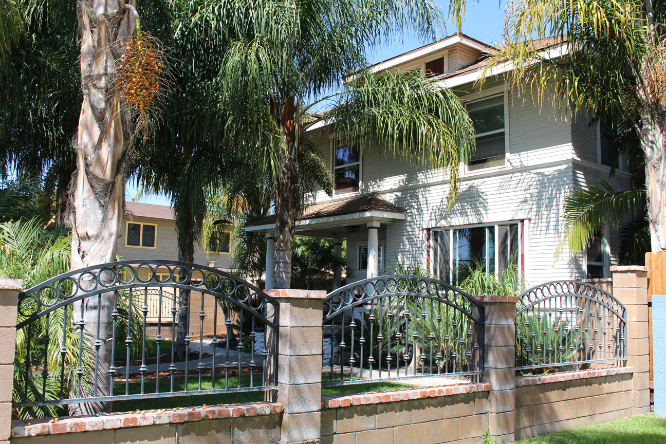 Two-story white residential building with mature palm trees, metal ornamental fencing, and green lawn from street view.