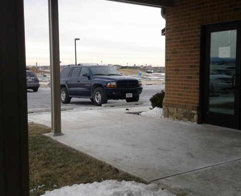 Exterior covered entryway with brick building facade, concrete pad, and parking area with vehicles.