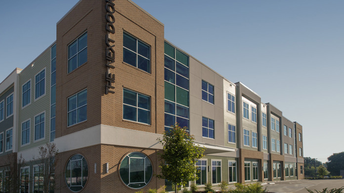 Exterior of modern treatment facility showing main entrance with 'The Next Door' signage, brick and metal panel construction, and large windows.