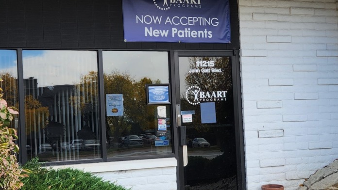 Exterior front entrance showing black door, white brick wall, landscaping, and 'Now Accepting New Patients' signage