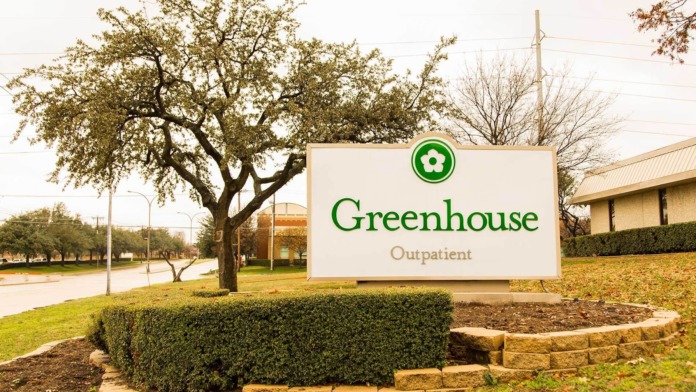Exterior showing Greenhouse Outpatient facility sign with green logo, landscaped grounds, mature trees, and stone retaining wall.