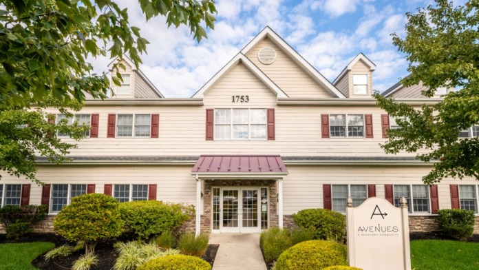 Exterior of two-story treatment center with cream siding, red shutters, landscaped grounds, and Avenues signage