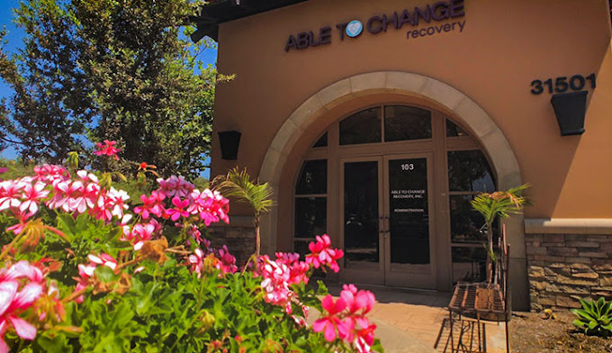 Exterior entrance of Able to Change Recovery showing tan stucco facade, arched doorway, facility signage, and flowering landscaping