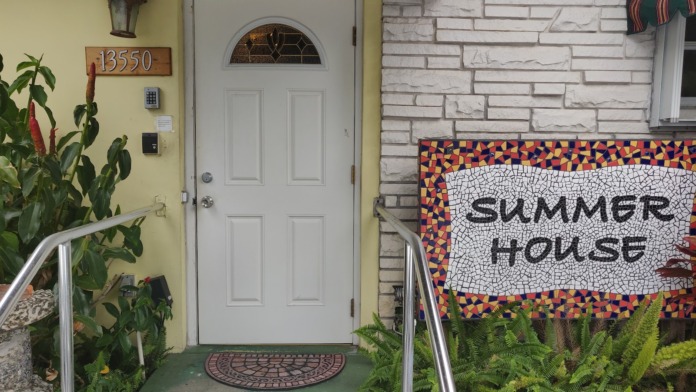 Exterior entrance showing white door, address 13550, colorful mosaic Summer House sign, and landscaping with tropical plants