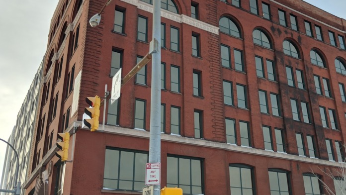 Exterior view of multi-story red brick building with regular windows, green awnings, and street-level entrance in urban setting.