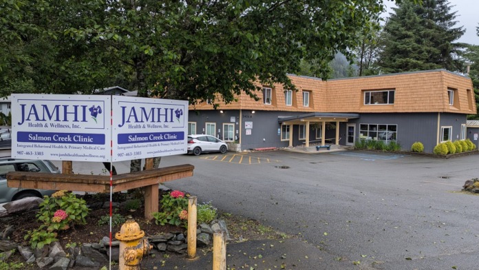 Exterior view of JAMHI treatment facility with wooden signage, brick building, parking lot, and evergreen landscape.