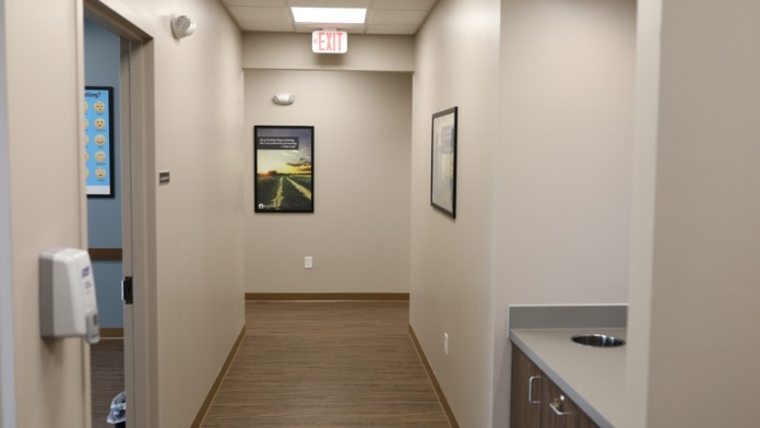 Interior hallway with exit signage, doors, framed artwork, white walls with wood trim, and fluorescent lighting