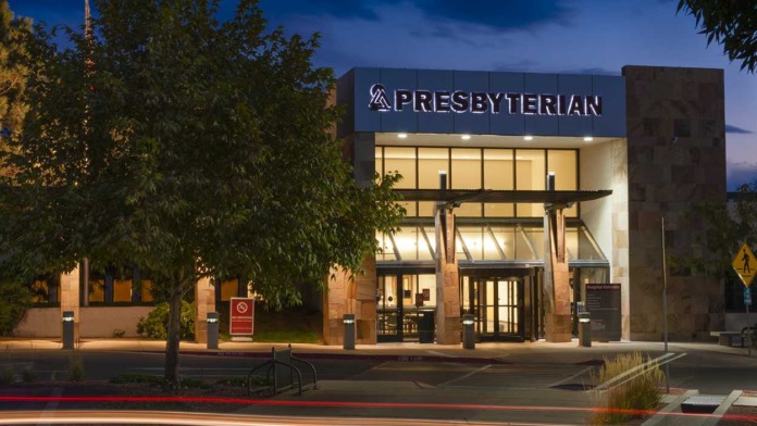 Exterior of main Presbyterian hospital building with illuminated signage, dark brick facade, large windows, and landscaping at twilight