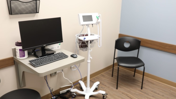 Interior clinical room with desktop computer setup, medical equipment stand with display monitor, chair, and flooring