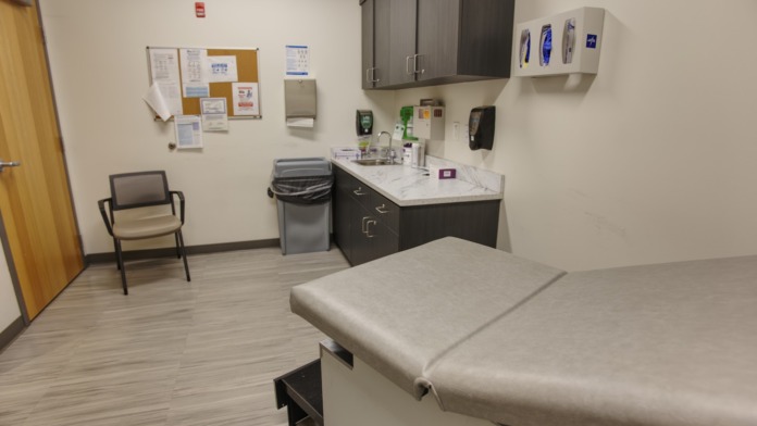 Interior clinical treatment room with examination table, medical sink, supply cabinet, bulletin board, and professional medical setup.